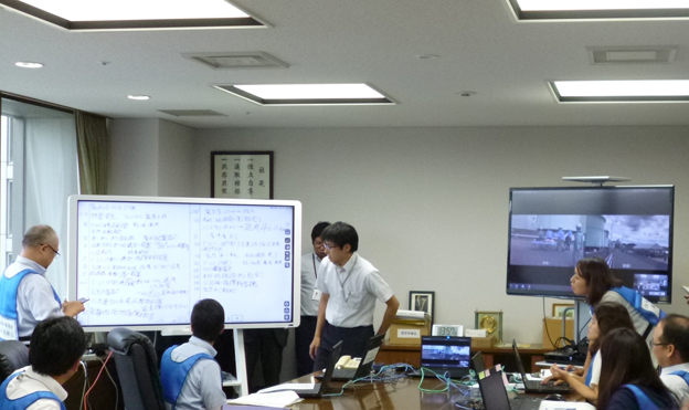 画像：東京本社に設置された対策本部。発災現場からリアルタイムに配信される映像（右側）と、四国事業所でIWB（左側）に集約した時系列の情報を元に、対策本部にて、現場への指示や様々な判断が行われている。