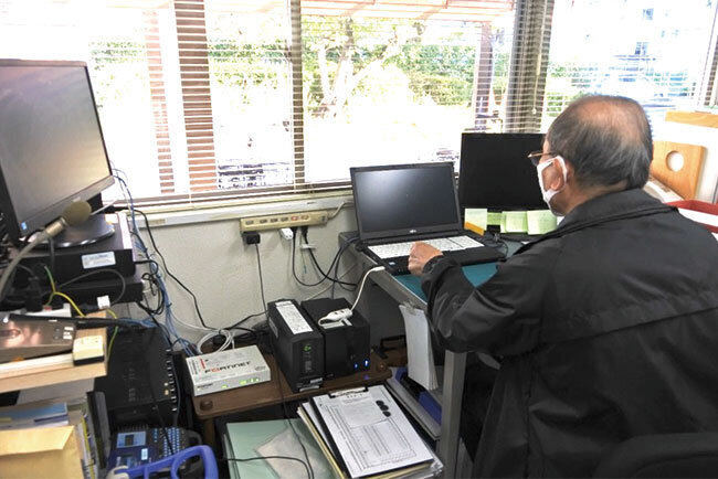 飯田さんの執務場所。机の左下に黒いNASと白いUTM機器が見える。左端に見えるのは駐車場監視カメラのモニター