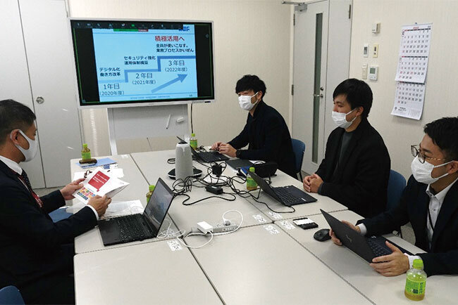 電子黒板を前にDXについて話し合うプロジェクトチームの今井担当部長（技術部）、長谷川係長（設備部）、五藤係長（総務部）、三上係長（設備部・リモート参加）（奥から）