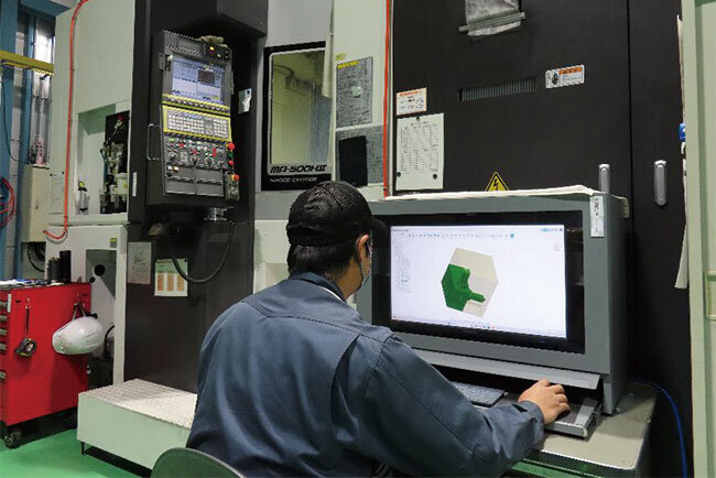 生産機械の前に置かれたパソコンでプログラム作成や3D画像確認をする従業員