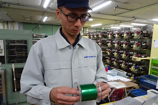 リールに巻かれたテープを手に「これよりさらに細いスリット加工もできます」と説明する製造部の大﨑亮祐課長＝越谷ワークス