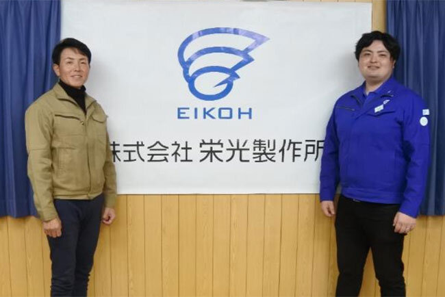 社旗の前に立つ勅使河原覚社長と勅使河原将也経営企画室室長