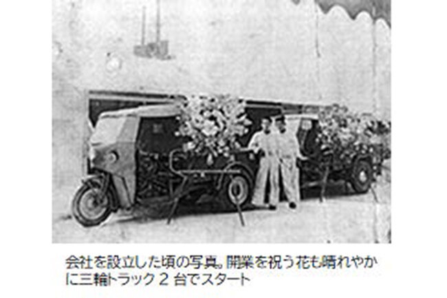会社を設立した頃の写真。開業を祝う花も晴れやかに三輪トラック2台でスタート