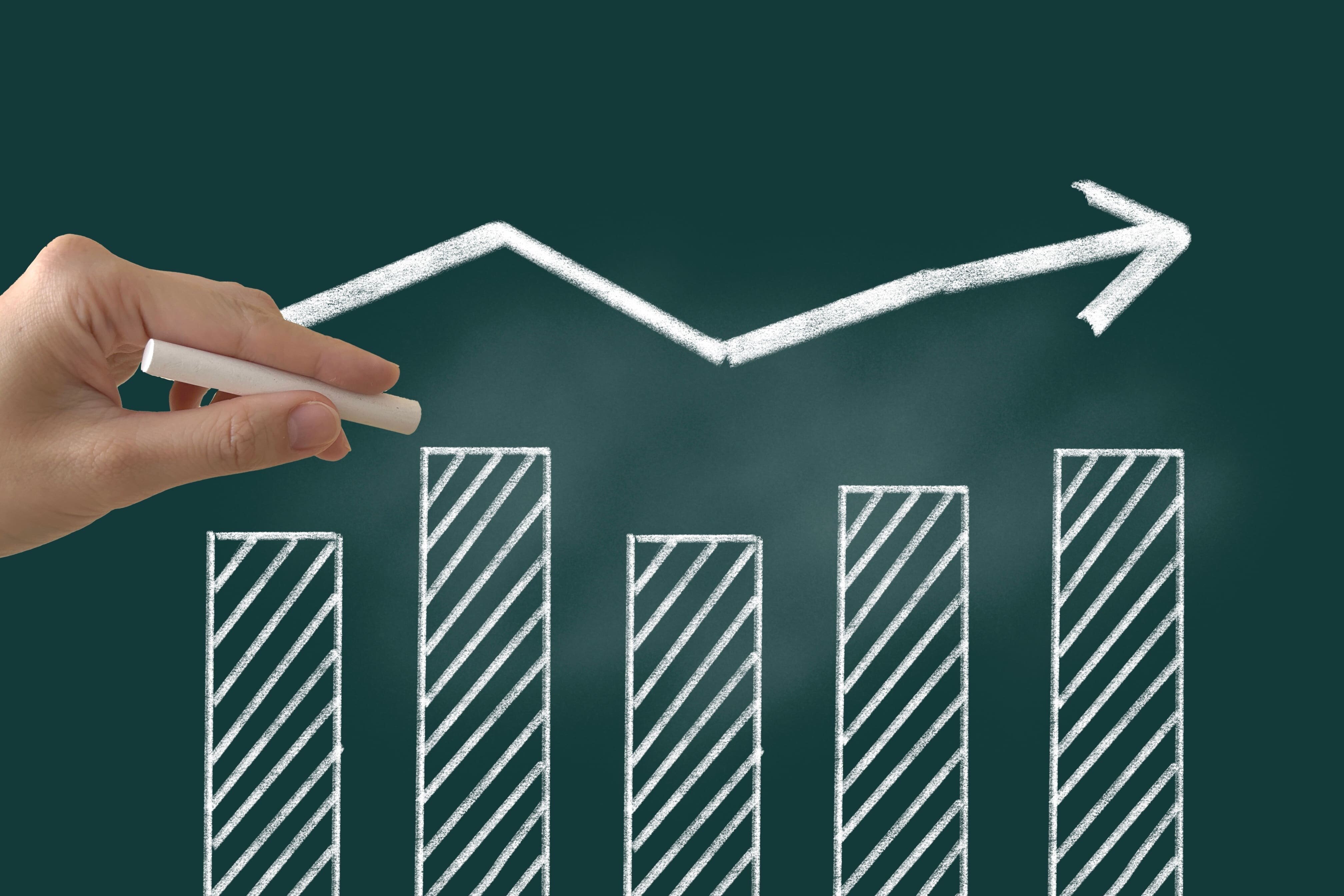 黒板にチョークで描かれた棒グラフと右上がりの矢印。手がチョークを持ち、成長や改善を示している。