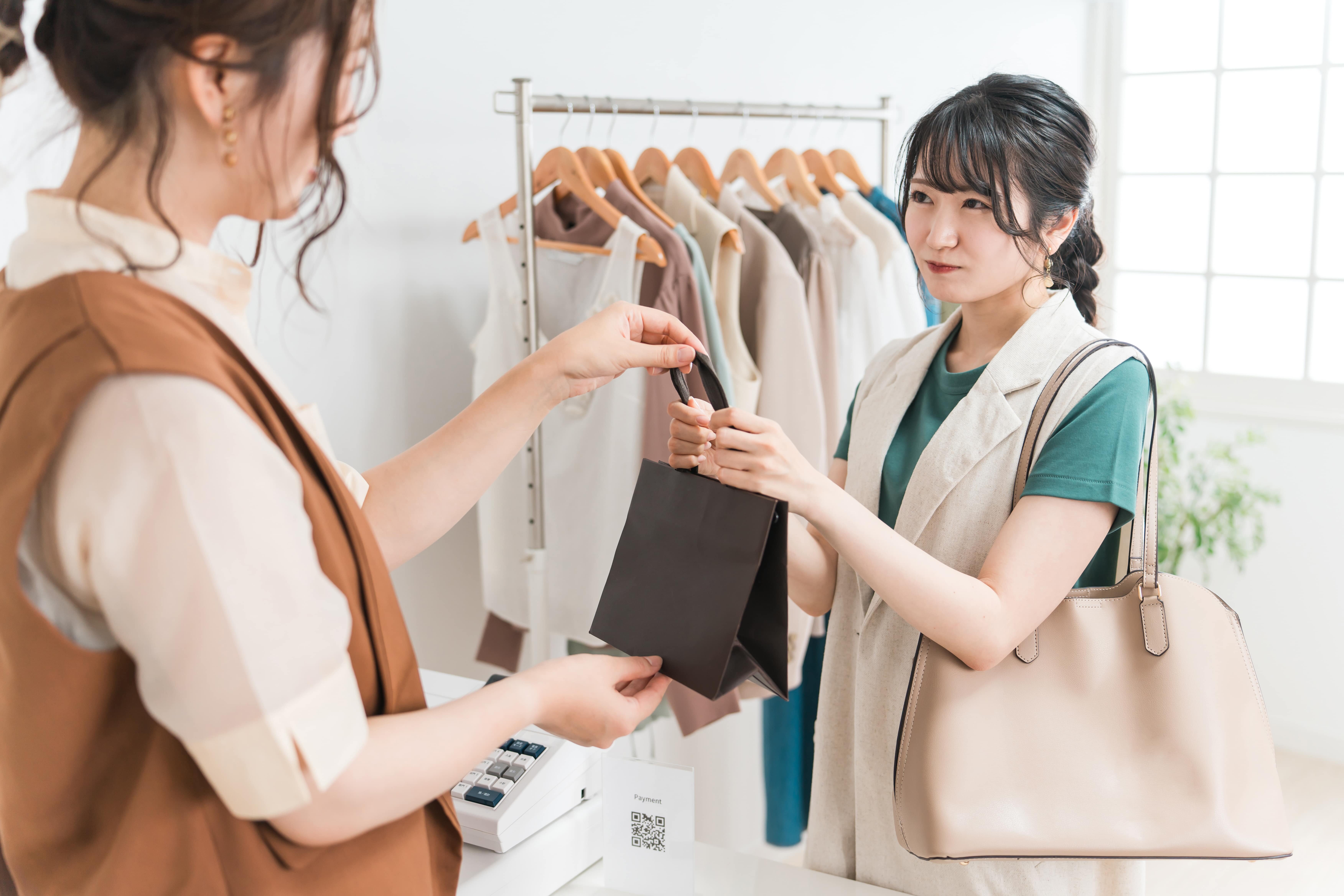 アパレルショップで店員から紙袋を受け取る女性の写真。 購入行動を通じて信頼や一貫性を形成する場面をイメージ。