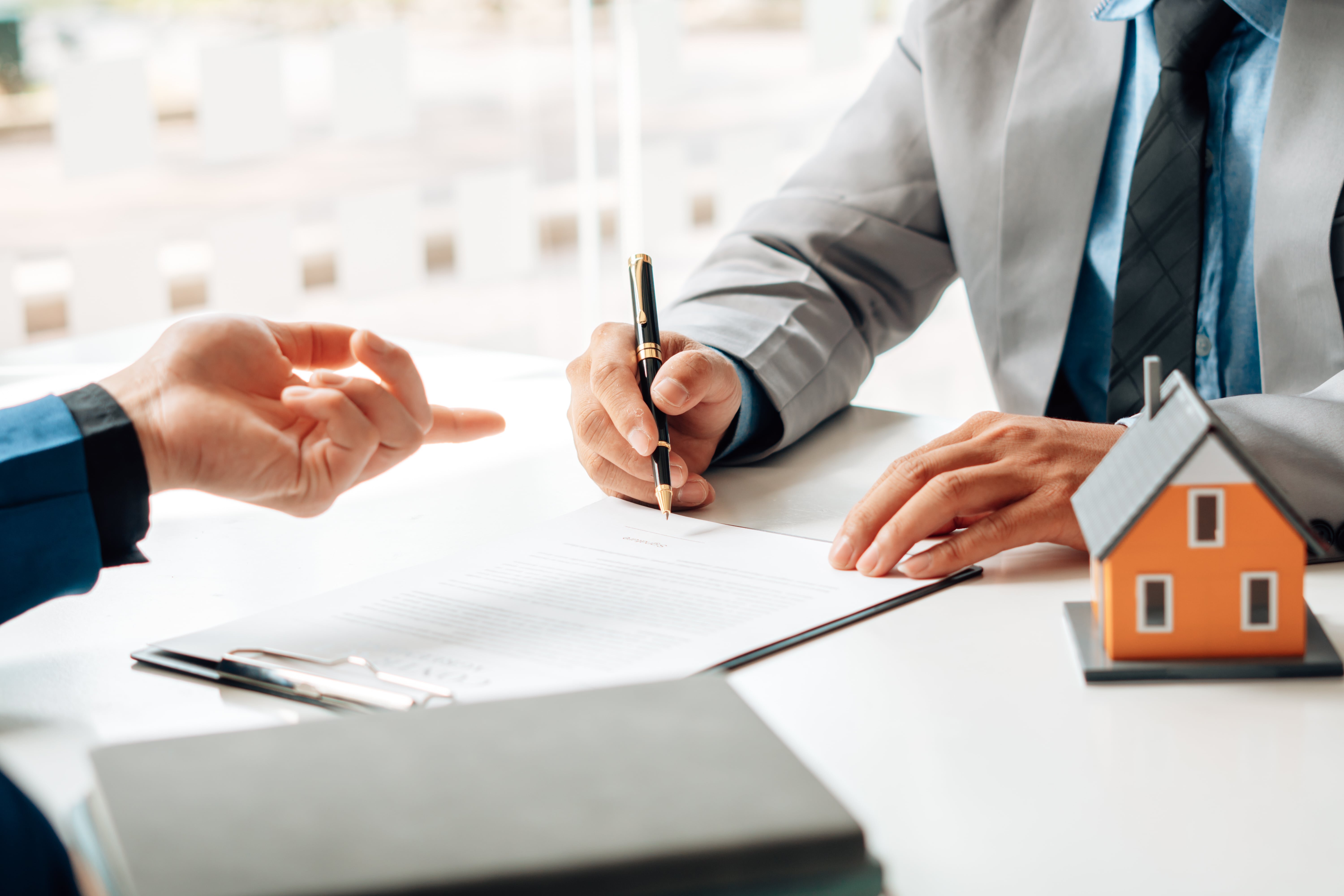 不動産契約書に署名する人と、家の模型を手に持つ人の手元を写した写真。契約や不動産取引のイメージを表している。