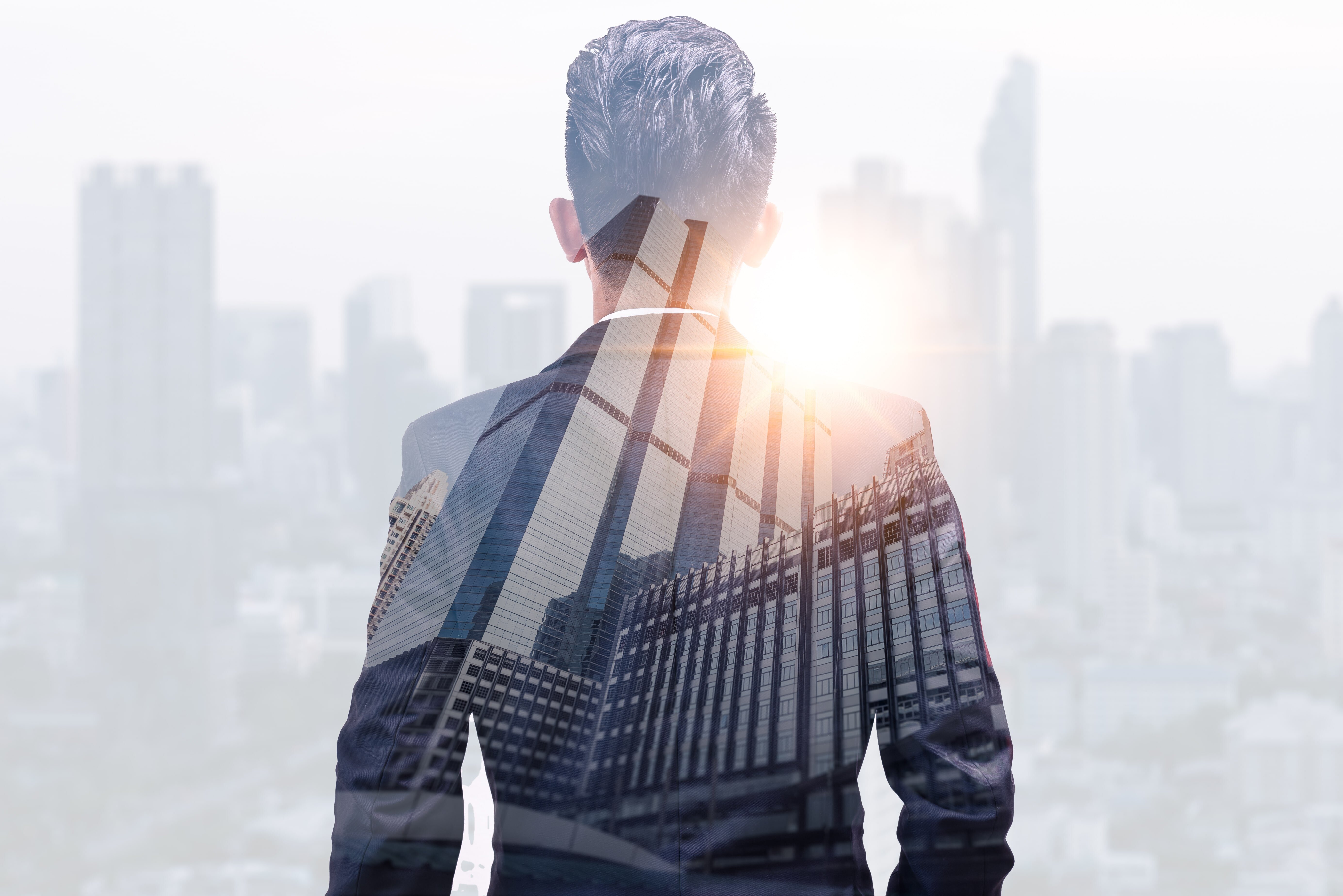 ビジネススーツ姿の男性が都市の高層ビル群を背景に立ち、朝日を受けて前を見据えている写真。 管理職としてのキャリアアップや成長、役職に就くことのメリットを象徴。